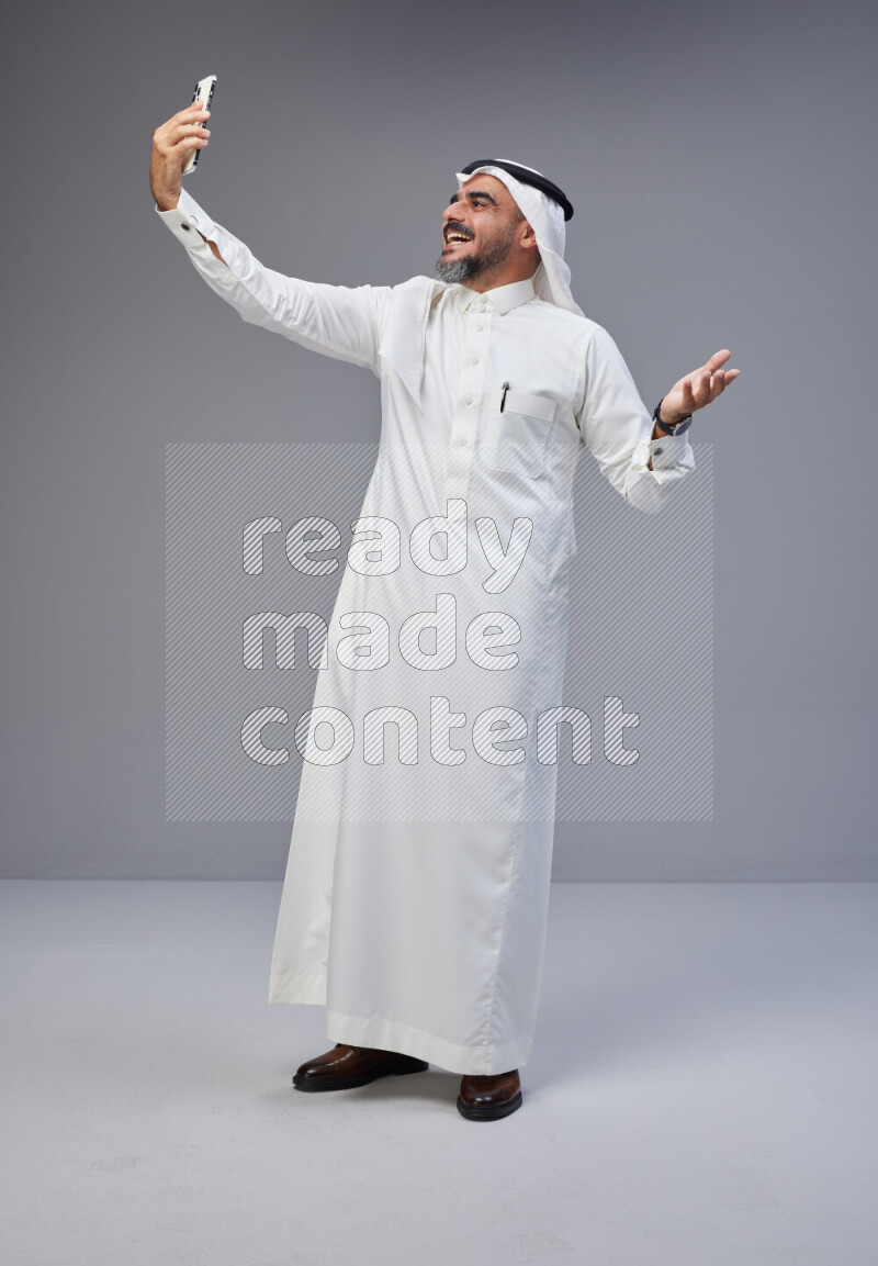 Saudi man Wearing Thob and white Shomag standing taking selfie on Gray background