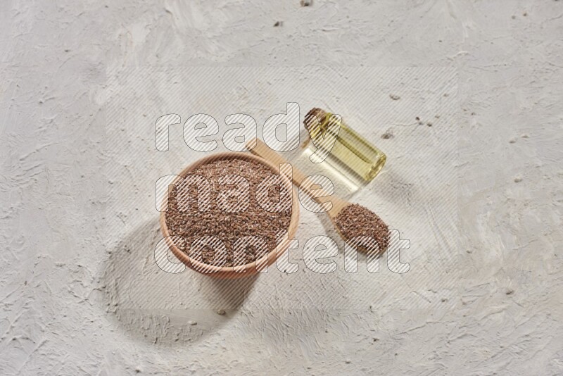 A wooden bowl and spoon full of flaxseeds with a bottle of flaxseeds oil on a textured white flooring