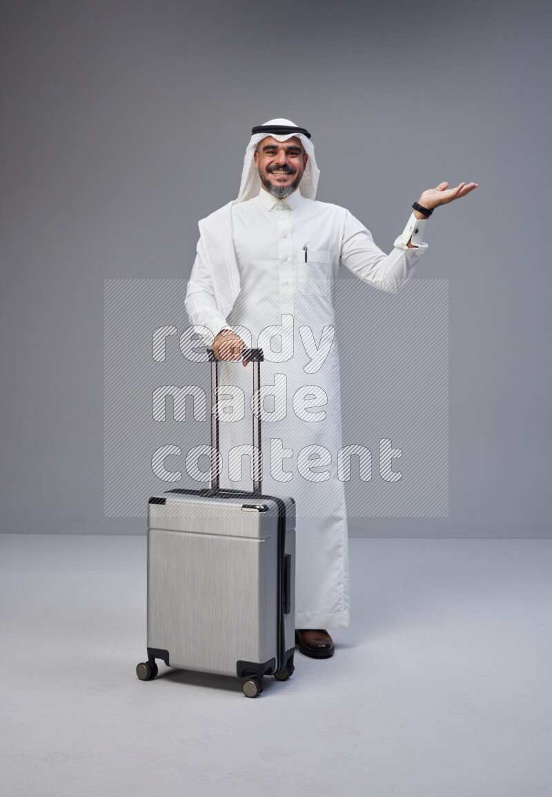 Saudi man wearing Thob and white Shomag standing holding Travel bag on Gray background