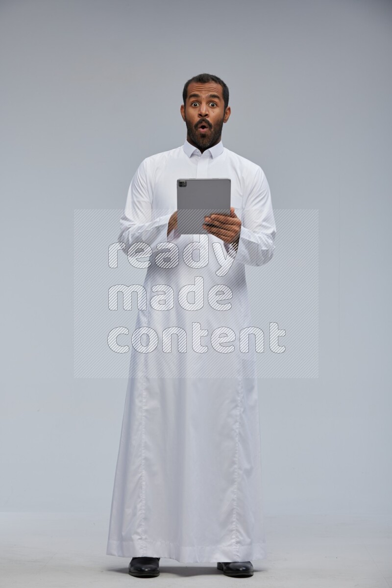 Saudi man wearing Thob standing working on tablet on Gray background