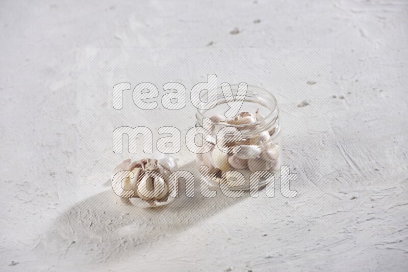 A glass jar full of garlic cloves with a whole garlic bulb beside it on a textured white flooring