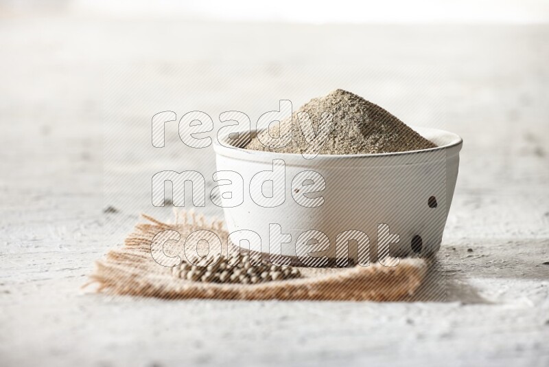 White pottery bowl full of white pepper powder set on a burlap piece of fabric with pepper beads and wooden pepper grinder on textured white flooring