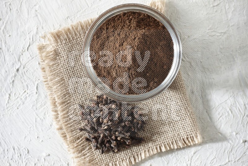 A glass bowl full of cloves powder and cloves grains on a burlap piece on a white flooring