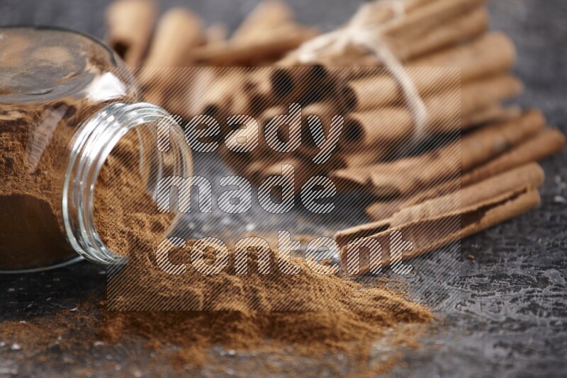 Herbal glass jar full of cinnamon powder flipped with cinnamon sticks stacked and bounded on a textured black background
