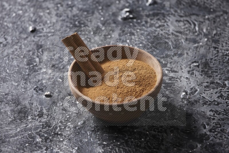 Wooden bowl full of cinnamon powder and a cinnamon stick on a textured black background