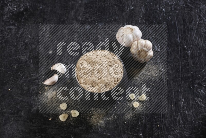 A black pottery bowl full of garlic powder with garlic cloves and a whole bulb beside it on a textured black flooring