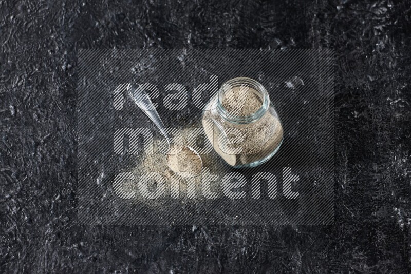 Herbal glass jar and metal spoon full of white pepper powder on textured black flooring