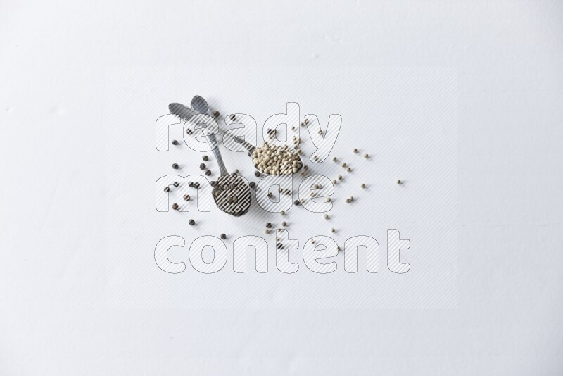 2 metal spoons full of black and white pepper beads with spreaded beads on white flooring