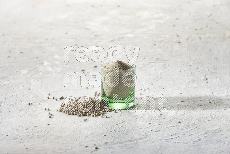 A green glass cup full of white pepper powder with white pepper beads on textured white flooring