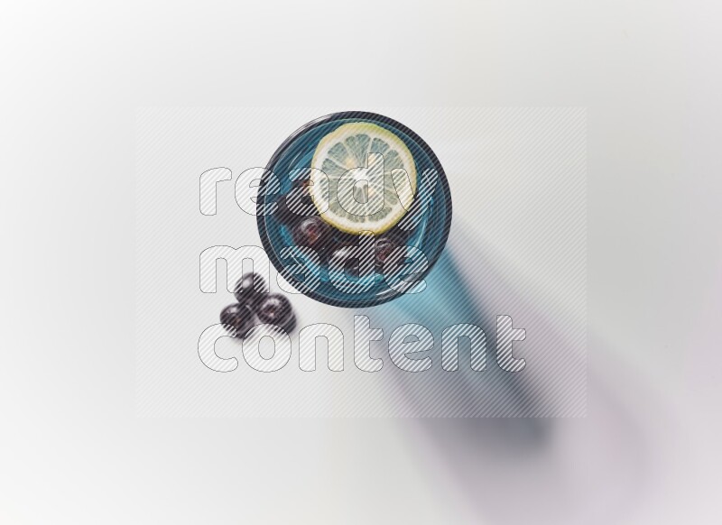 Blue transparent glass filled with blueberries decorated with lemon slices on a white background