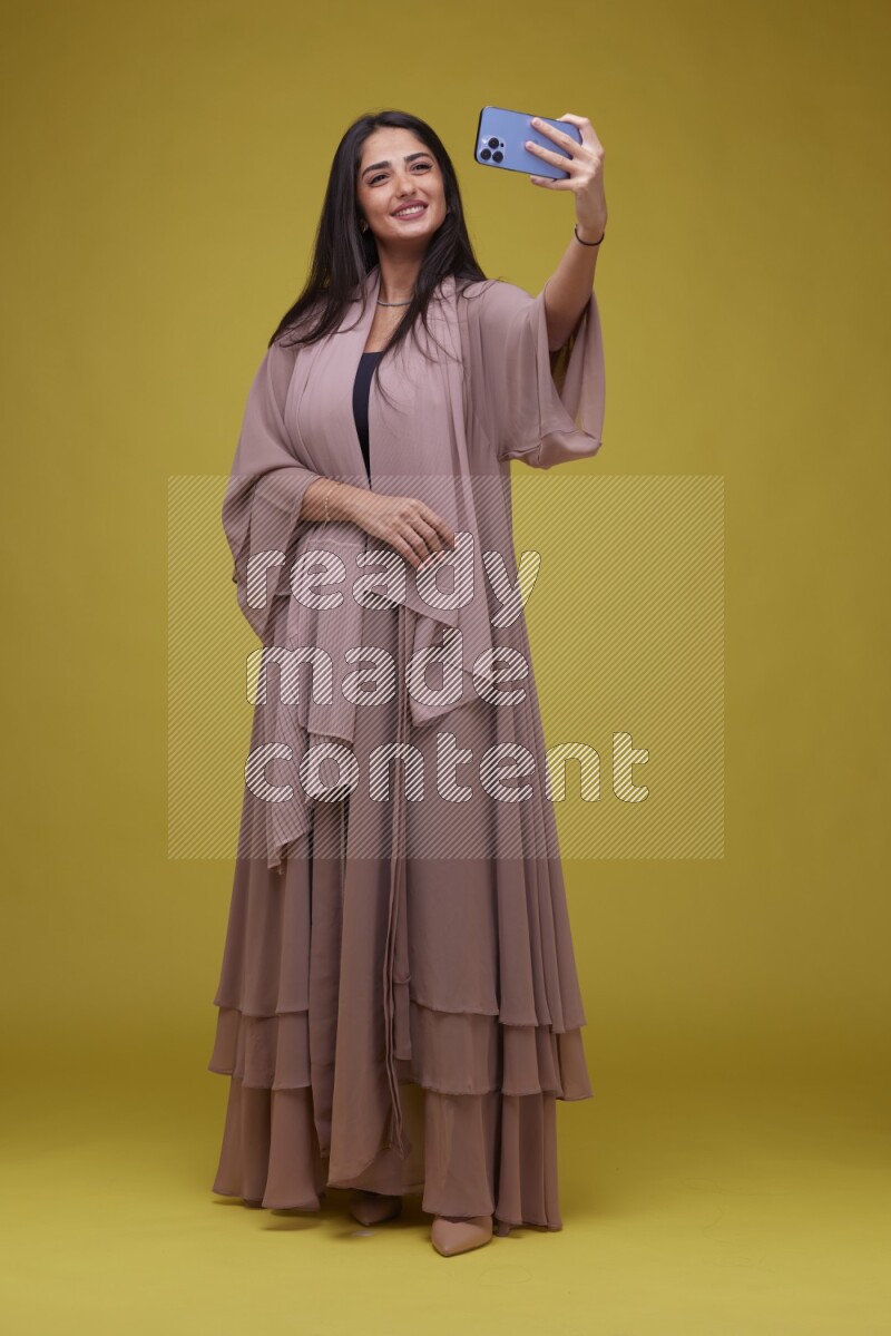 A woman Taking a Selfie on a Yellow Background wearing Brown Abaya