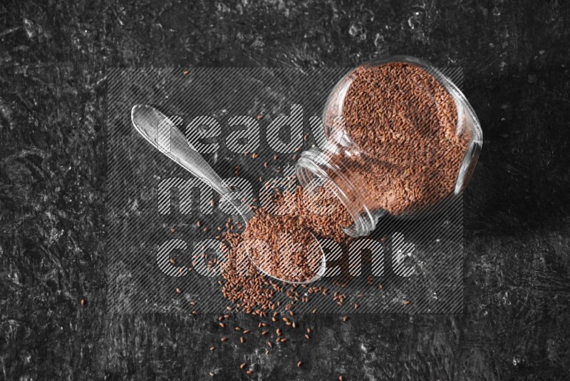 A glass spice jar full of garden cress seeds flipped and seeds spread out with a metal spoon full of the seeds on a textured black flooring