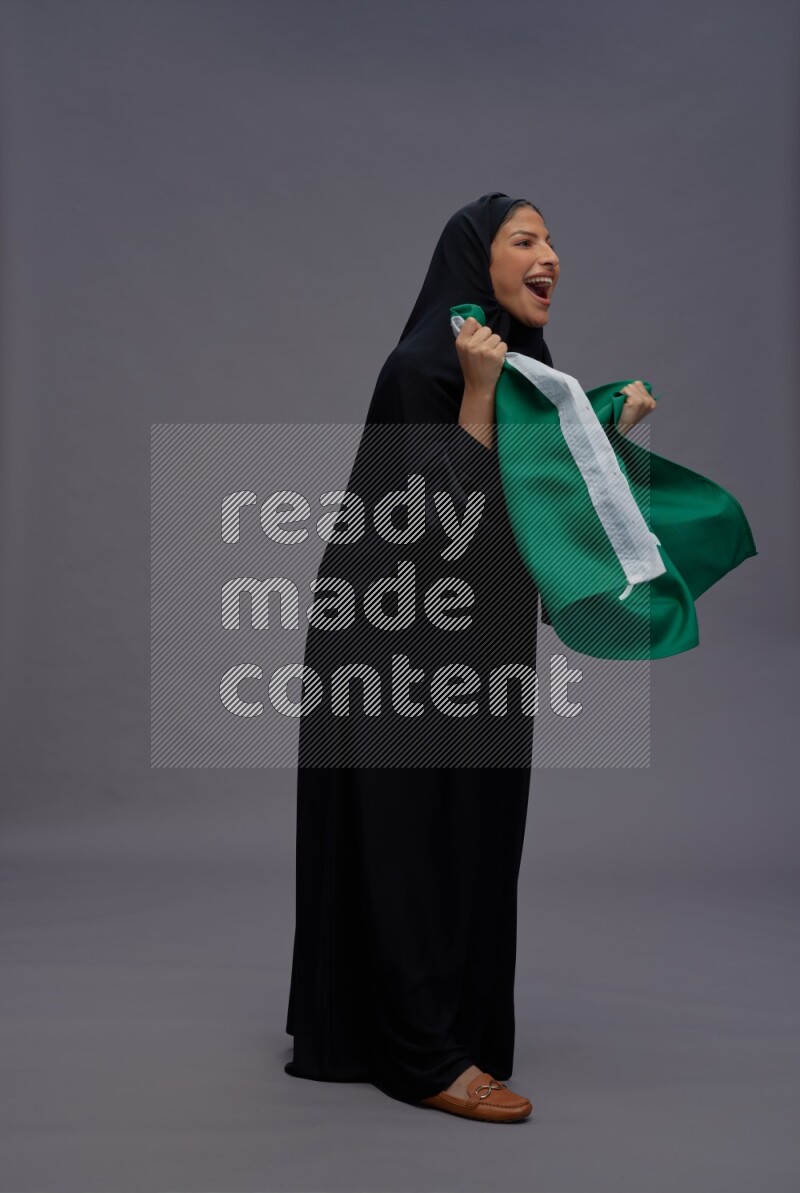 Saudi woman wearing Abaya standing holding Saudi flag on gray background