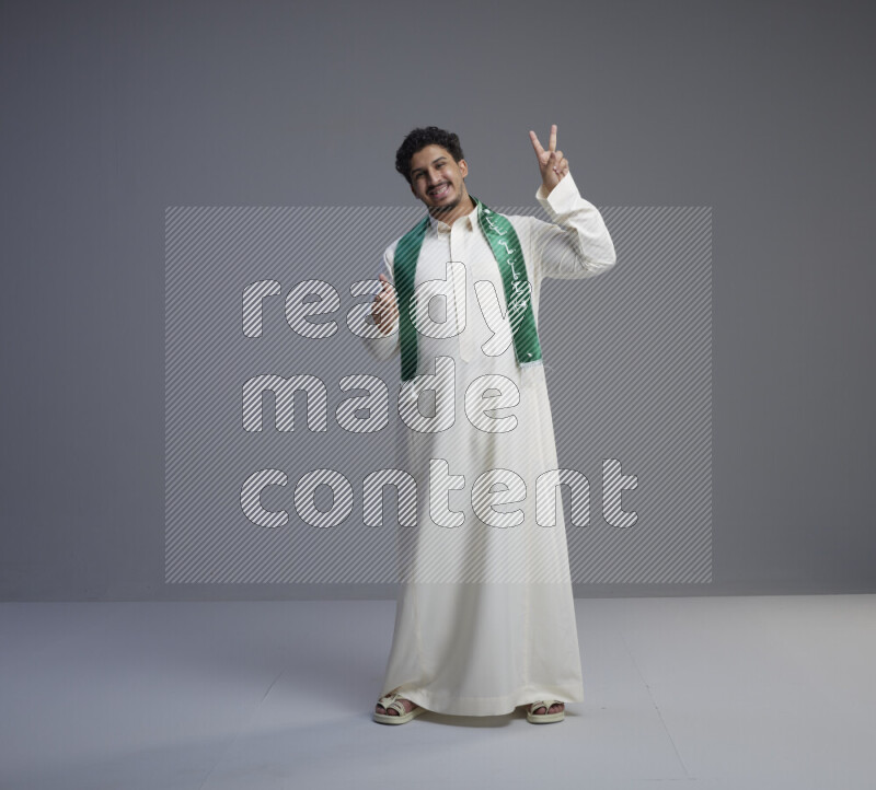 A Saudi man standing wearing thob and Saudi flag scarf on gray background