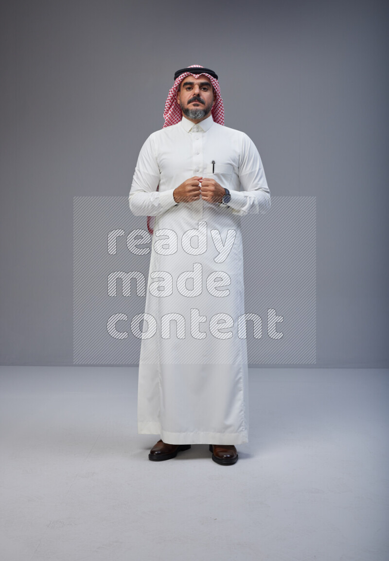 Saudi man Wearing Thob and red white Shomag standing interacting with the camera on Gray background