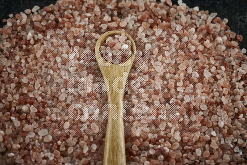 A wooden spoon full of coarse himalayan salt crystals on a bunch of the crystals on black background