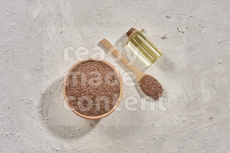 A wooden bowl and spoon full of flaxseeds with a bottle of flaxseeds oil on a textured white flooring