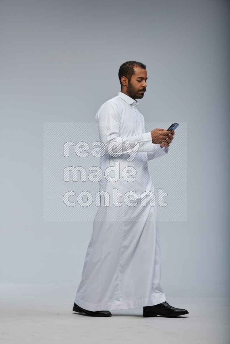 Saudi man wearing Thob standing texting on phone on Gray background