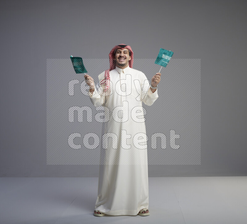 A Saudi man standing wearing thob and red shomag raising small Saudi flag on gray background
