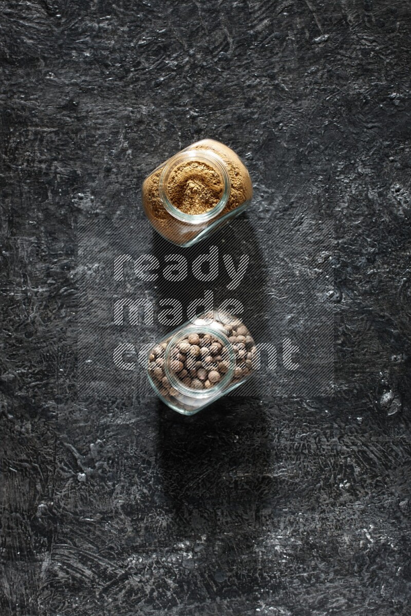 2 glass spice jars full of allspice powder and whole balls on a textured black flooring