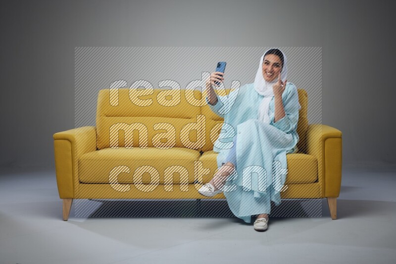 A Saudi woman wearing a light blue Abaya and white head scarf sitting on a yellow sofa and taking selfies with her phone eye level on a grey background