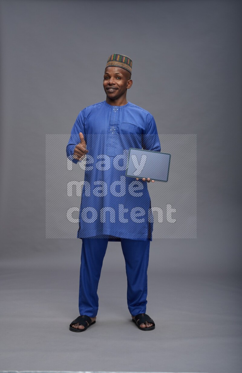 Man wearing Nigerian outfit standing showing tablet to camera on gray background