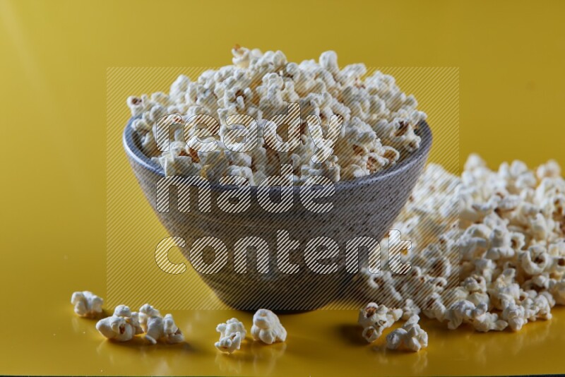 A multicolored pottery bowl full of popcorn with popcorn beside it on a yellow background in different angles
