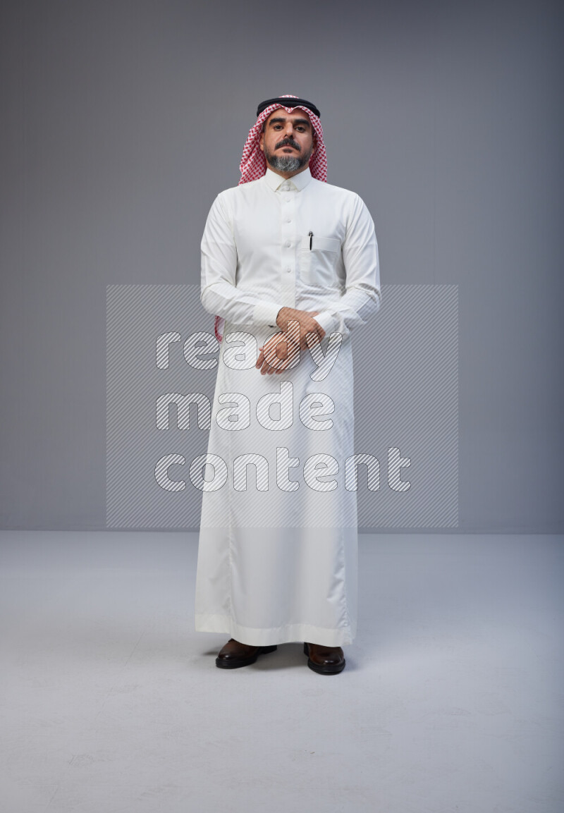 Saudi man Wearing Thob and red white Shomag standing interacting with the camera on Gray background