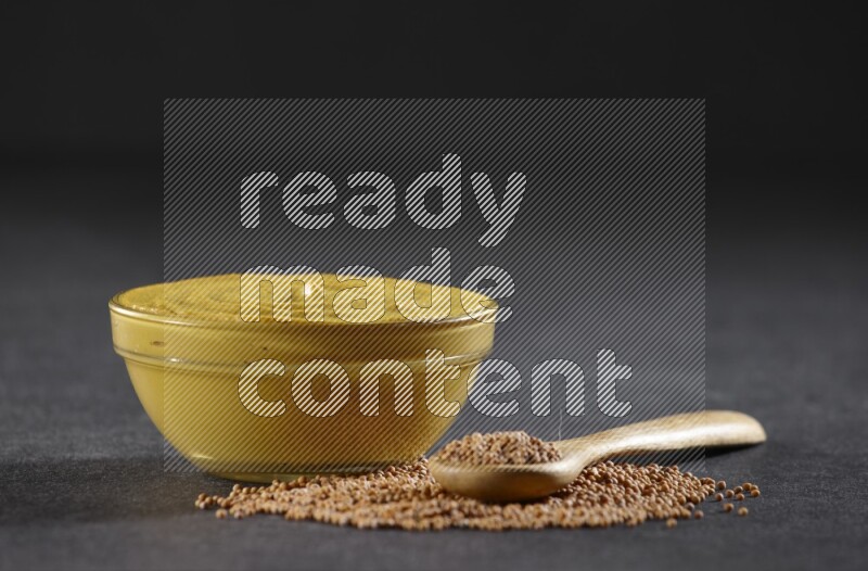 A glass bowl full of mustard paste with mustard seeds underneath and a full wooden spoon on black flooring