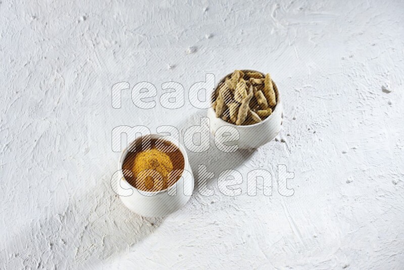 2 beige pottery bowl full of turmeric powder and dried turmeric whole fingers on textured white flooring