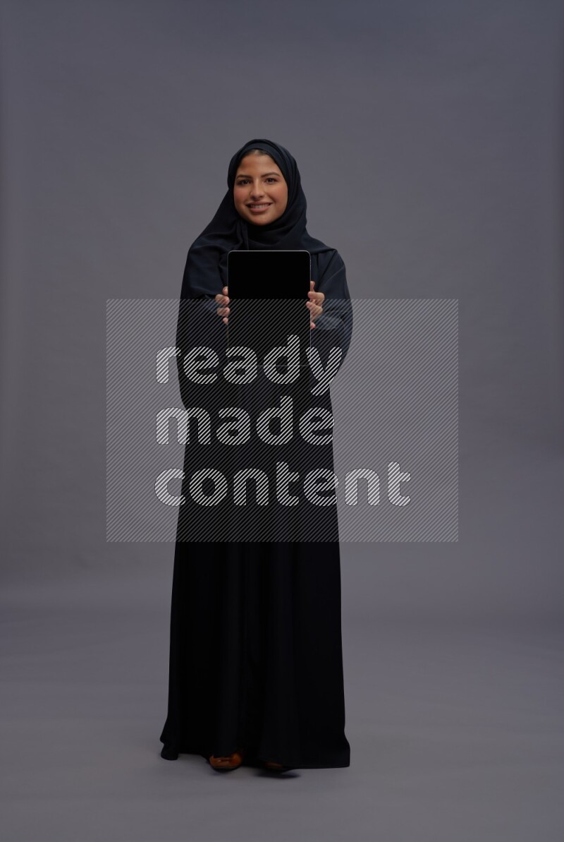 Saudi woman wearing Abaya standing showing tablet to camera on gray background