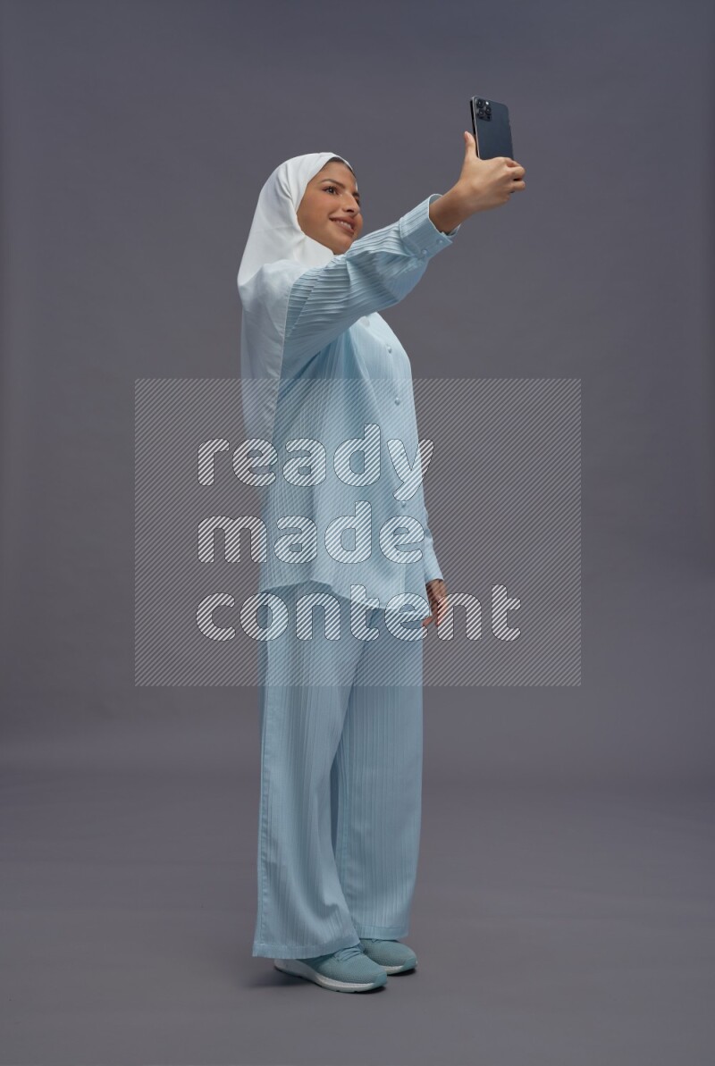 Saudi woman wearing hijab clothes standing taking selfie on gray background