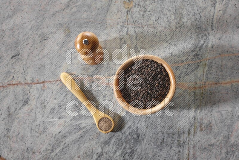 A wooden bowl full of black pepper and a wooden spoon full of black pepper powder and a wooden grinder on a marble flooring