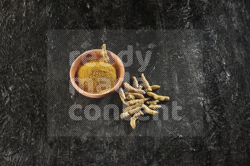 A wooden bowl full of turmeric powder with dried turmeric whole fingers on textured black flooring