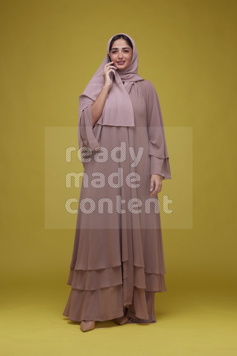A woman having a call on a Yellow Background wearing Brown Abaya with Hijab
