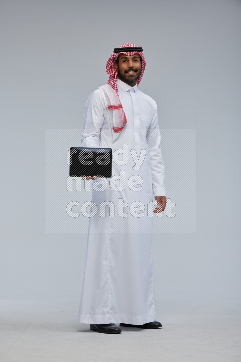 Saudi man Wearing Thob and shomag standing showing tablet to camera on Gray background