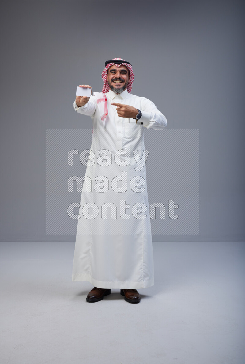 Saudi man Wearing Thob and red Shomag standing holding ATM card on Gray background