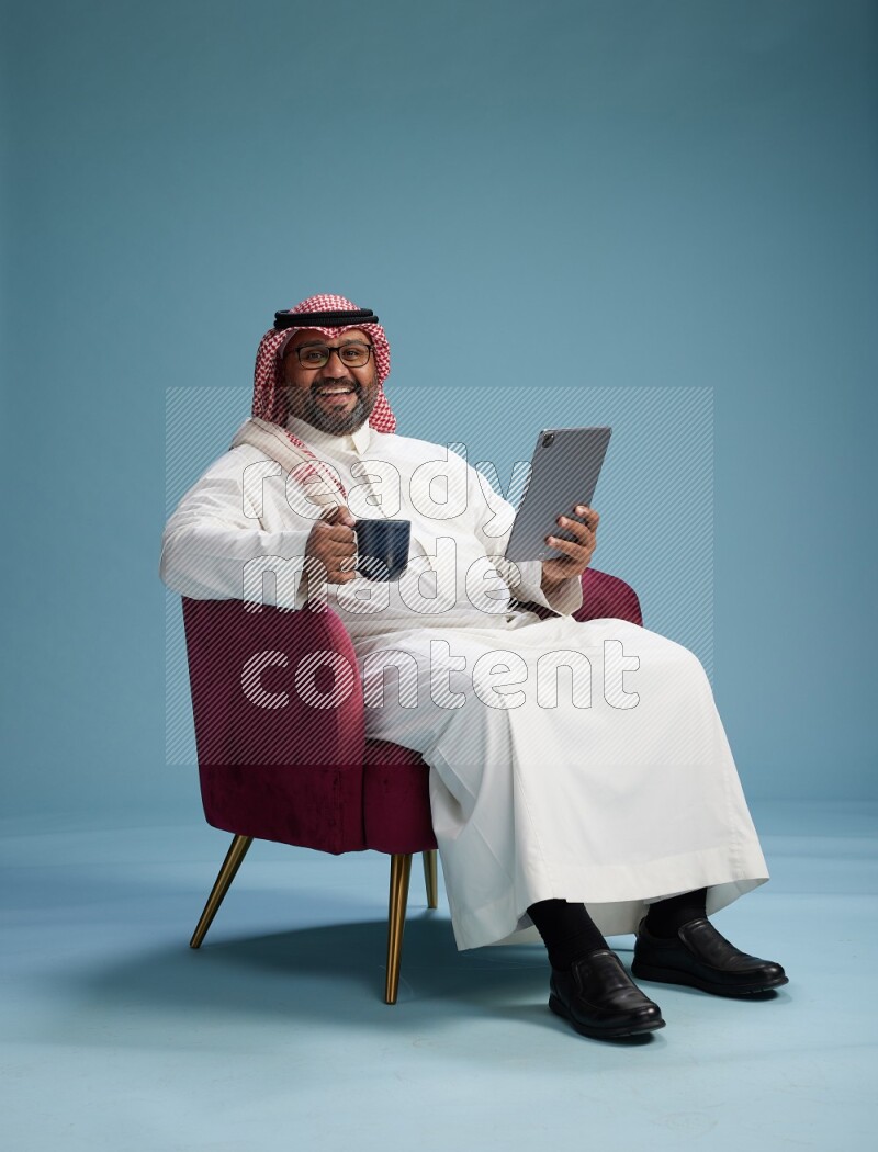 Saudi Man with shimag sitting on chair working on tablet on blue background