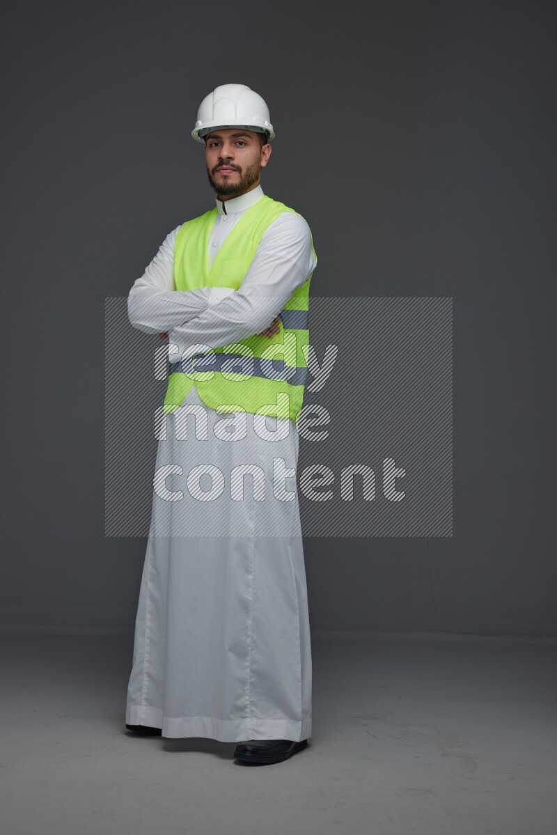 A Saudi man wearing Thobe with a yellow safety vest and white helmet standing and crossing his hands eye level on a gray background