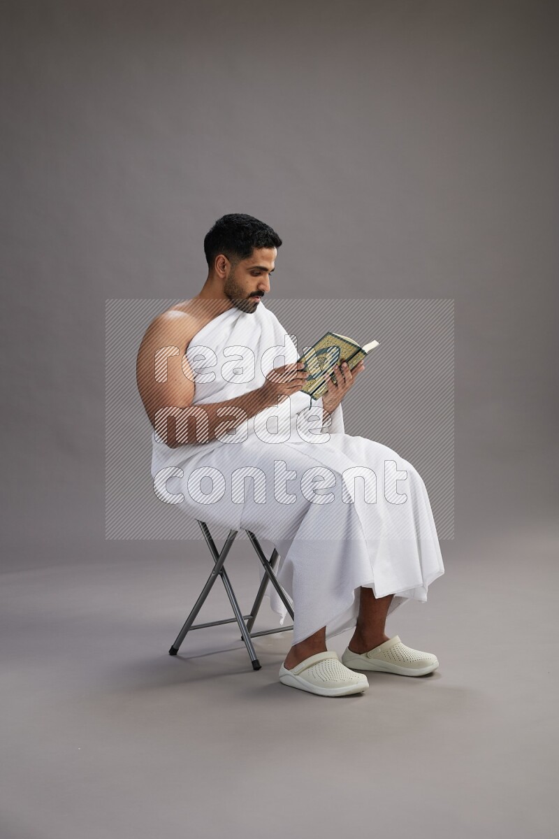 A man wearing Ehram sitting on chair reading quran on gray background