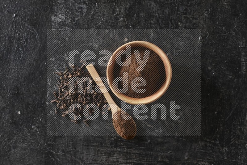 A wooden bowl and a wooden spoon full of cloves powder with spreaded cloves on a textured black flooring