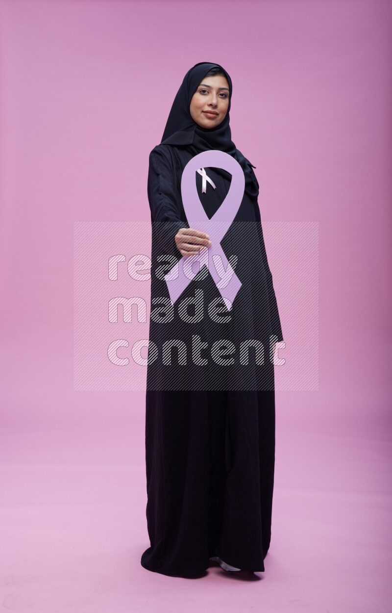 Saudi woman wearing pink ribbon on abaya standing holding awareness ribbon on plain pink background