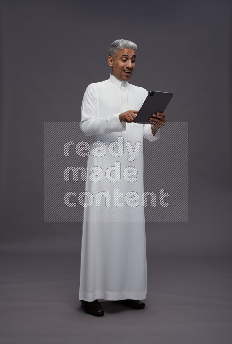 Saudi man wearing thob standing working on tablet on gray background