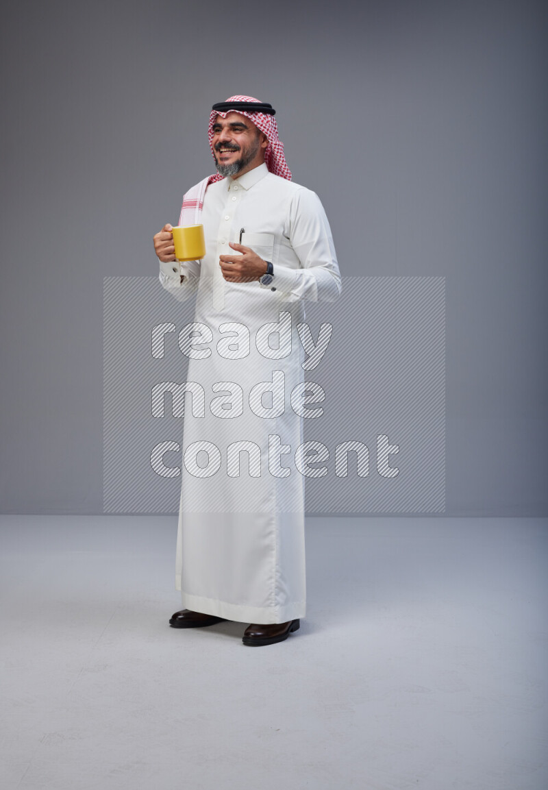 Saudi man Wearing Thob and red Shomag standing holding mug on Gray background