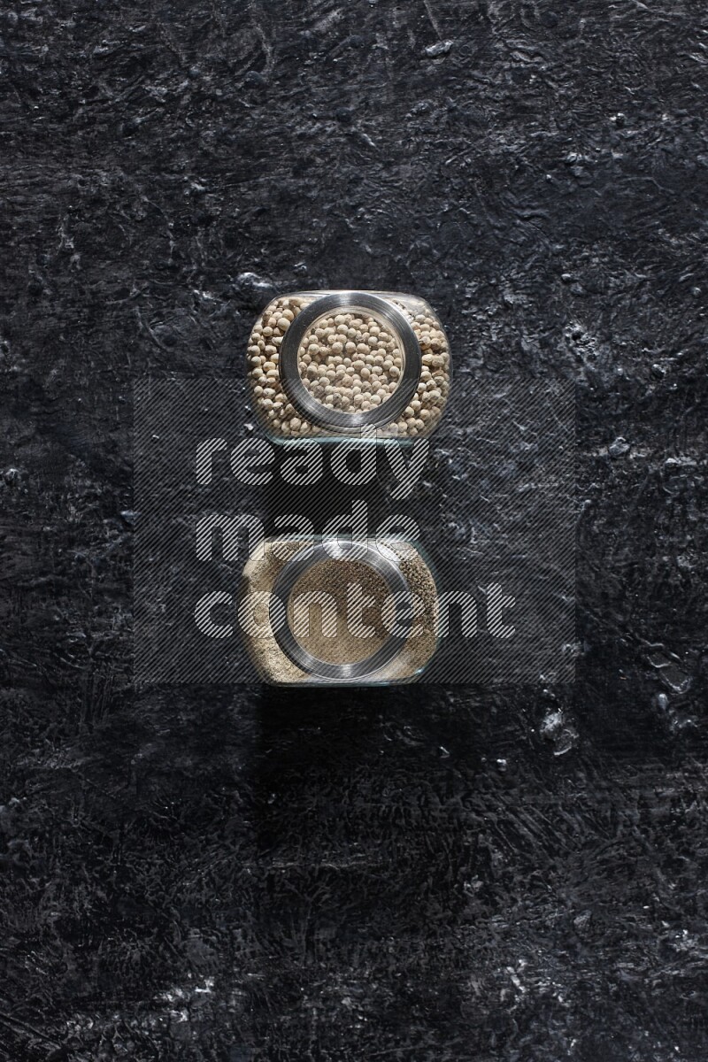 2 Herbal glass jars full of white pepper beads and powder on textured black flooring