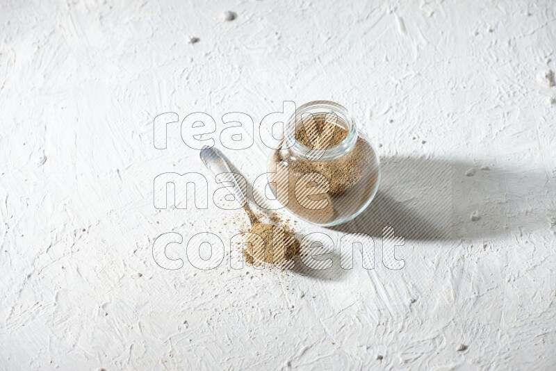 A glass spice jar and metal spoon full of cumin powder on textured white flooring