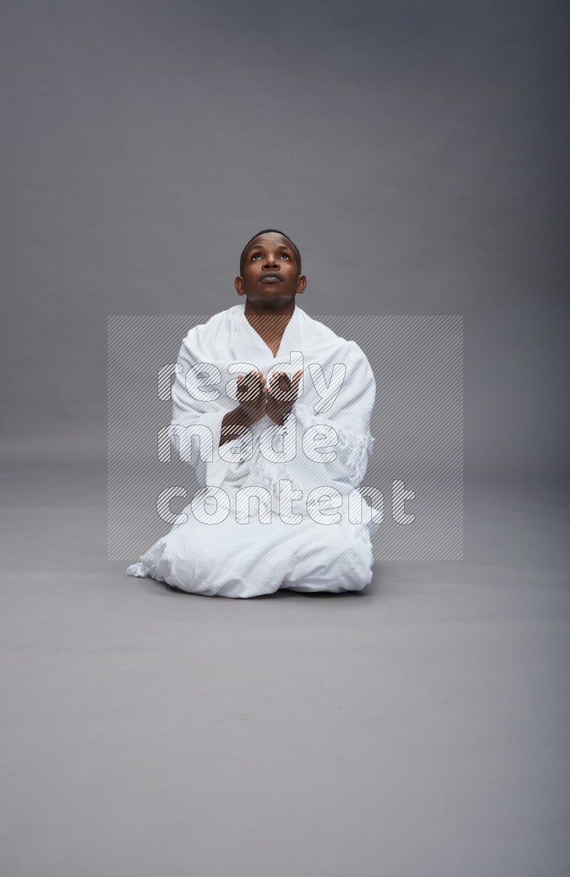 A man wearing Ehram sitting on floor dua'a on gray background