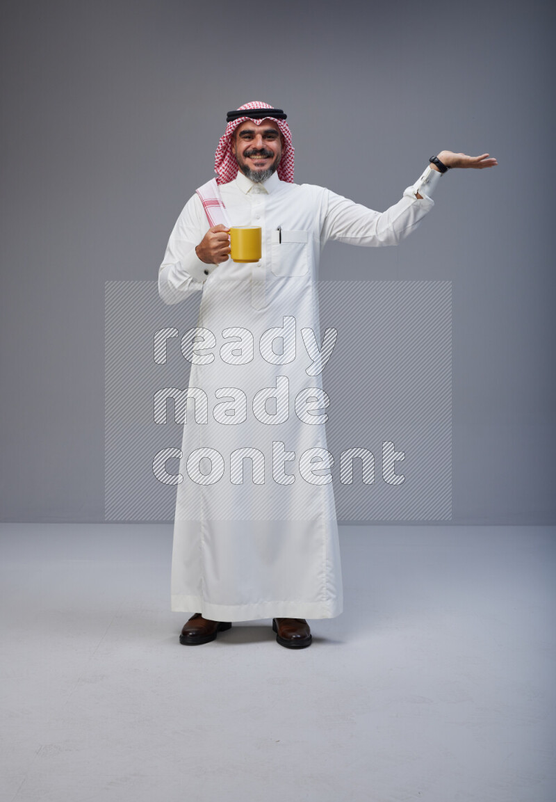 Saudi man Wearing Thob and red Shomag standing holding mug on Gray background