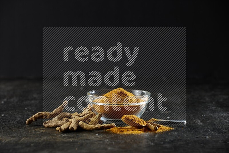 A glass bowl and a metal spoon full of turmeric powder and dried whole fingers next of them on textured black flooring