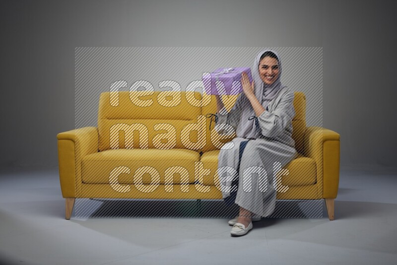 A Saudi woman wearing a light gray Abaya and head scarf sitting on a yellow sofa holding a gift box beside multi color gift boxes eye level on a grey background